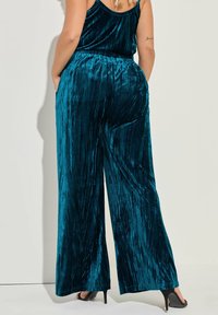 Teal fluwelen wijde broek met een gestructureerde, geribde afwerking en elastische tailleband. Beschikt over een vloeiende silhouet en is gecombineerd met zwarte hakken.