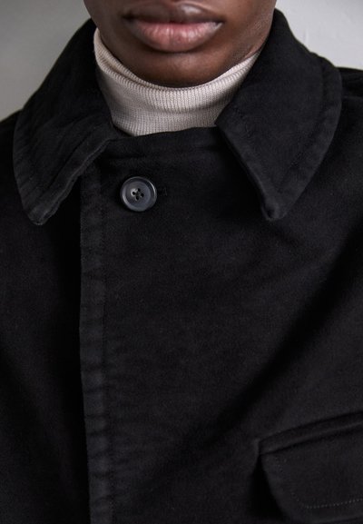 Black wool coat with a large collar, featuring a single button closure. Worn over a light turtleneck. Soft texture and tailored fit.