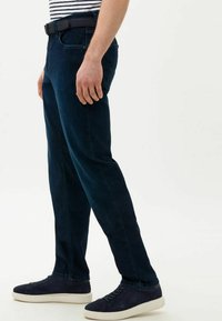 Donkerblauwe spijkerbroek met een rechte pasvorm, voorzien van een riem, voorzakken en subtiele stiksels. Draagt met marineblauwe sneakers.