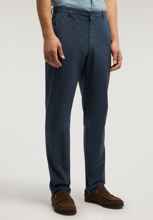 Uomo che indossa pantaloni blu navy e mocassini scamosciati marroni, in piedi contro uno sfondo chiaro e uniforme.