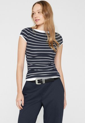 Junge Frau trägt ein tailliertes kurzärmeliges Oberteil mit marineblauen und weißen Streifen, dazu marineblaue Hose und einen schwarzen Gürtel, blickt zur Seite.