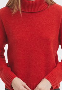 Frau trägt einen leuchtend roten Strickrollkragenpullover mit langen Ärmeln und hält ihre Hände nahe der Taille zusammen.