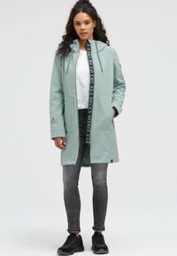 Light green waterproof coat with a zip closure, front pockets, and a hood. Features a contrasting text detail on the zipper and sleeves.