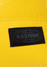 Gul vattentät material med synliga vattendroppar, med en svart tygpatch som visar "EASTPAK"-logotypen och jord globdesign.
