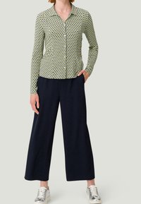 Langarm, knoopbare shirt in een groen en zwart geometrisch patroon, gecombineerd met een broek met wijde pijpen in marineblauw en zilveren sneakers.