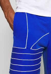 Modré sportovní šortky s bílými horizontálními a zakřivenými pruhy, obsahující boční kapsu a měkkou, strukturovanou tkaninu.