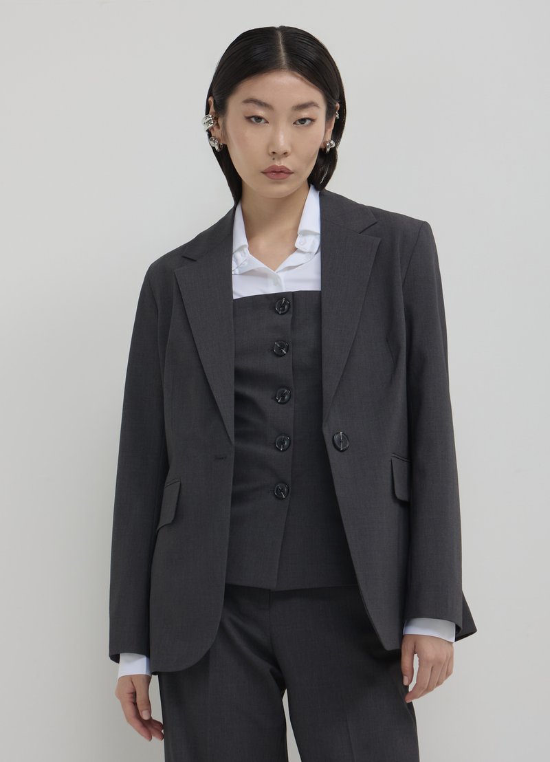 Blazer gris ajusté avec revers en pointe, deux poches frontales et une fermeture à un bouton, associé à un gilet assorti et une chemise blanche en dessous.