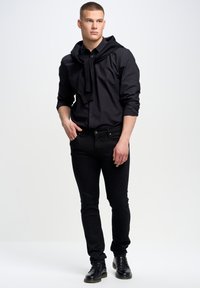 Camisa negra de botones con mangas largas y una capucha texturizada caída en el cuello, combinada con jeans negros ajustados y botas negras con cordones.