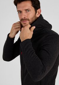 Dunkelgrauer Reißverschluss-Hoodie aus strukturiertem Stoff, mit hohem Kragen, langen Ärmeln und einem Frontreißverschluss, mit einem kleinen roten Akzent.