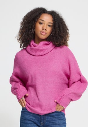 Femme aux cheveux bouclés portant un pull à col roulé rose avec des boutons aux poignets et un jean bleu, debout les mains dans les poches.