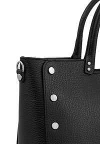 Black textured leather handbag corner with silver round studs and double handles against white background.
