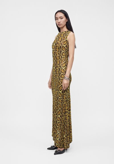 Femme debout portant une robe sans manches, longue jusqu'au sol, jaune et marron à motif serpent, avec des chaussures noires pointues et un bracelet argenté.
