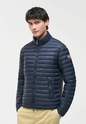 Giovane uomo che indossa una giacca trapuntata blu navy con le mani in tasca e pantaloni beige contro uno sfondo chiaro e semplice.