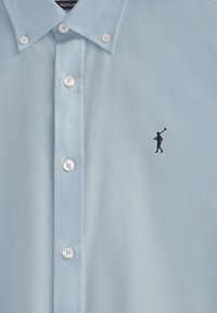 Ljusblå skjorta med krage gjord av mjukt tyg, med vita knappar och en svart broderad logga av en golfspelare på vänster sida.