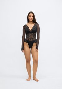 Črno čipkasto bodi z lahkimi dolgimi rokavi, globokim V-izrezom in zapletenimi cvetličnimi vzorci. Hlačke z visokim izrezom z detajli čipkastega roba.