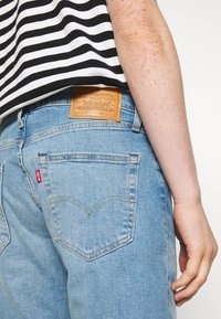 Vaaleansinisiä farkkuja, joissa on merkitty nahkapaikka takana, vakiotaskut takana ja erottuva ompelu.