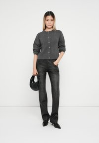 Cárdigan gris de punto con cuello redondo y cierre de botones, combinado con jeans negros acampanados. La modelo lleva un bolso negro y calza zapatos de punta.