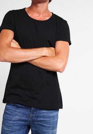 Uomo con le braccia incrociate che indossa una maglietta nera a maniche corte e jeans blu su uno sfondo bianco uniforme.