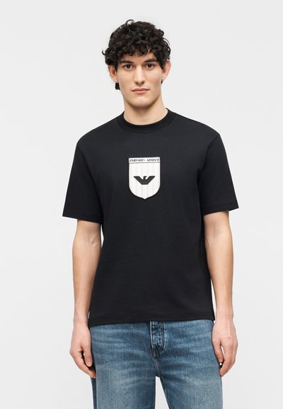 Νέος άνδρας με σγουρά μαλλιά φοράει μαύρο t-shirt Emporio Armani με κεντημένη ετικέτα λογότυπου και μπλε τζιν, στέκεται μπροστά σε λευκό φόντο.