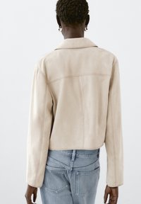 Beige mockaskinnkort jacka med snibbig krage, synliga sömmar och långa ärmar, tillsammans med ljusblå denimjeans.