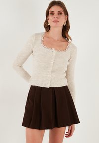 Kreminė megzta palaidinė su kvadratiniu iškirpimu ir nėriniuotu apvadu, priekyje su šešiais mygtukais, derinama su rudomis plisuotomis sijonais.