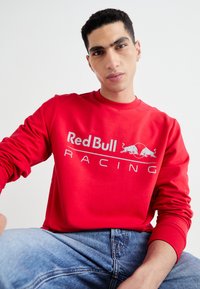 Sudadera de crewneck roja en algodón brillante con un logo blanco de "Red Bull Racing" y un gráfico de dos toros. Combinada con jeans de mezclilla azul.