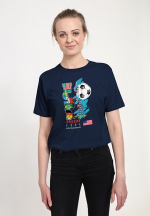 Kvinde iført en mørkeblå T-shirt med farverig tekst "World Cup Soccer 1994", fodboldgrafik og amerikansk flag, kombineret med sorte bukser.