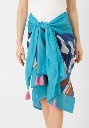 Vrouw draagt een blauw sarong met patroon, in de taille gestrikt met roze kwastjes, gecombineerd met een wit mouwloos top, tegen een witte achtergrond.