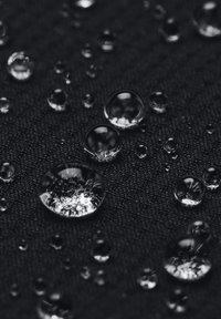 Gocce d'acqua su una superficie nera texturizzata, che mostrano varie dimensioni e forme, con una finitura lucida che riflette la luce.