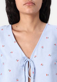 Blouse rayée bleu et blanc à manches courtes, ornée d'une petite broderie de cerises rouges et d'un lien au niveau du col. Texture de tissu lisse.