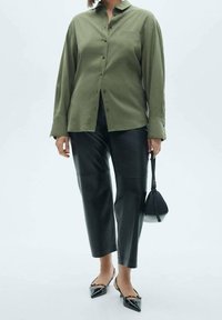 Chemise boutonnée vert olive à manches longues, associée à un pantalon en cuir noir fuselé et à des escarpins noirs à bouts pointus. Petit sac à main noir.