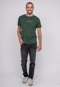 T-shirt vert à manches courtes arborant le texte "BLUE & Jeans" dans un design multicolore texturé. Porté avec un jean foncé et des baskets blanches.