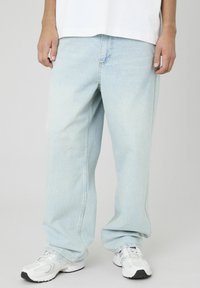 Jean en denim oversize bleu clair avec une coupe droite et des détails de couture visibles, assortis avec des chaussures de sport blanches.