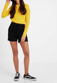 Žena v žlutém háčkovaném topu s dlouhým rukávem, černých šortkách s vysokým pasem a tkaničkou, a černobílých sportovních teniskách, stojící na bílé pozadí.