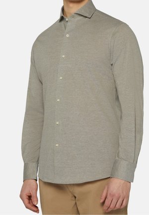 Mand iført en lysegrå langærmet skjorte med krave og beige bukser mod en ensfarvet baggrund.