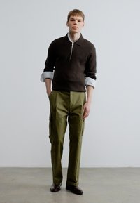 Brūns pulovers ar rāvējslēdzēju, uzvilkts pār gaiši zilu kreklu ar apkakli, kopā ar olīvu zaļiem darba biksēm un tumši brūnā apavos. Minimālistisks dizains.