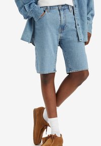 Pantalones cortos de mezclilla azul claro con un corte relajado, que cuentan con una cinturilla y bolsillos frontales, combinados con zapatos de ante marrón y calcetines blancos.