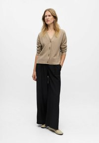 Beige cardigan with a v-neck and button closure, combined with loose black pants, and light shoes, set against a neutral background.