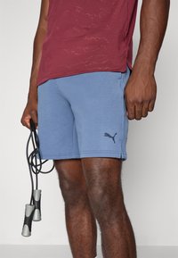 Puma sportshorts in een blauwe katoenmix met een zwart logo. Het model houdt een zwarte springtouw vast met zilverkleurige handvatten en draagt een bordeauxrood shirt.