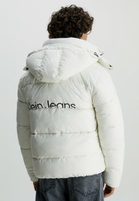 Bílá péřová bunda s kapucí, která má prošívané části a černé logo "Klein Jeans" na zádech. Volný střih a hladká textura.