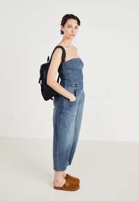 Jeansbyxdress med axelbandslös design, ljusblå färg, framfickor och smalnande ben. Stylistiskt parat med bruna mockaskor och en svart ryggsäck.
