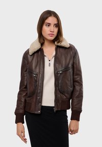 Veste bombardier en cuir marron avec un col en shearling, fermeture éclair sur le devant, deux grandes poches et des poignets côtelés. Apparence texturée et chaude.