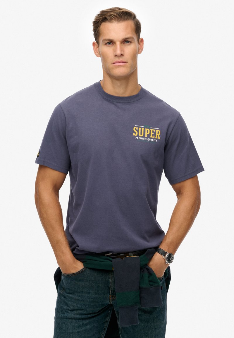 Donkerblauwe T-shirt met ronde hals en een vooropdruk met de tekst "SUPER PREMIUM QUALITY." Gecombineerd met denim en een groen-navy gestreepte trui om de taille.