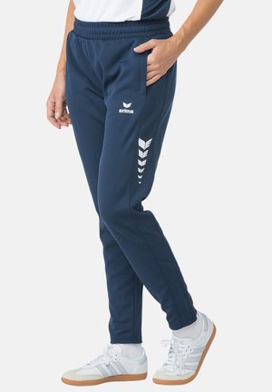 Person, die navyblaue Erima-Trainingshose und weiße Adidas-Sneaker mit Gummisohlen trägt, steht mit einer Hand in der Hosentasche vor einem einfarbigen Hintergrund.