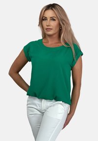 Zielona koszulka z krótkim rękawem, okrągłym dekoltem i miękką fakturą, zestawiona z białymi dopasowanymi spodniami. Materiał wydaje się lekki.