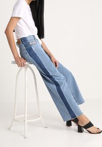 Svetle modré kalhoty s širokými nohavicemi a tmavě modrými bočními pruhy, s vysokým pasem a rovný střih. Nosené s bílým zkráceným topem a černými block heels.