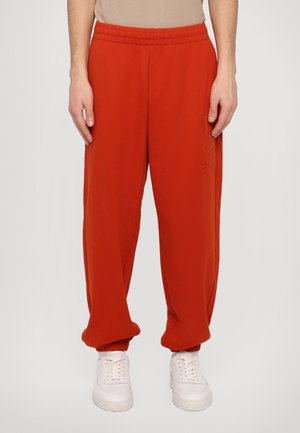Pantalons de survêtement rouges avec une taille élastique et des poignets, ornés d'un subtil logo sur le côté, associés à des baskets blanches.