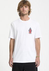 Hvid bomulds T-shirt med et lille, farverigt grafik på venstre bryst, der viser en rød figur udskåret mod trekantede former.