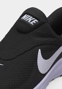 Μαύρο αθλητικό παπούτσι Nike slip-on με λευκό λογότυπο Swoosh και υφασμάτινο πάνω μέρος με υφή, που δείχνει τη μαξιλαρωμένη γλώσσα με ανάγλυφο το λογότυπο Nike.