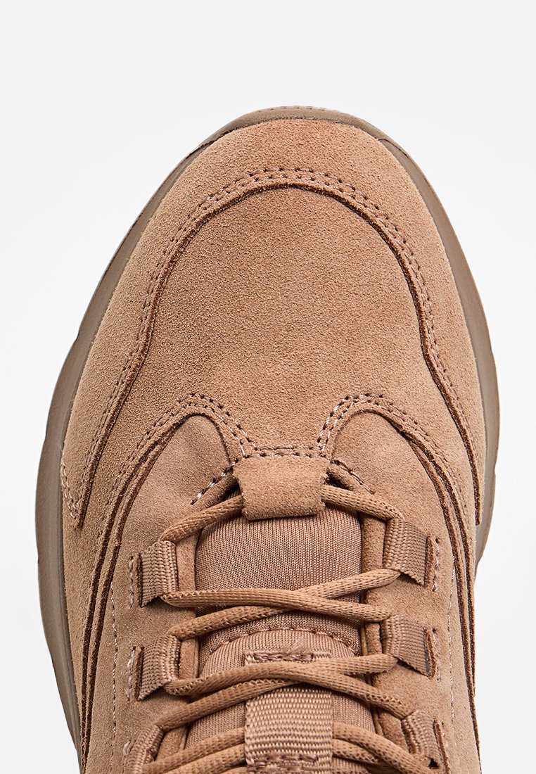 Zapatilla de ante marrón con puntera redondeada, superficie texturizada y detalles de cordones. Cuenta con costuras en contraste y un cuello acolchado para mayor comodidad.